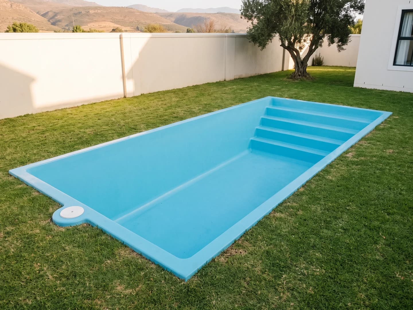 Piscina de fibra de vidrio de 10,00 por 4,00 metros instalada a nivel del pasto en el jardín de una casa, vista desde arriba, vacía y sin borde, con la aleta de fibra a la vista, la tapa redonda del skimmer en un costado y la escalera de cuatro peldaños a lo ancho de un extremo