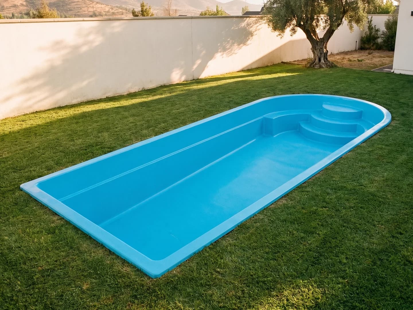 Piscina de fibra de vidrio de 10,50 por 4,00 metros instalada a nivel del pasto en el jardín de una casa, vista desde arriba, vacía y sin borde, con la aleta de fibra a la vista y el extremo redondeado con una banca curva y una escalera de tres peldaños en abanico en la esquina