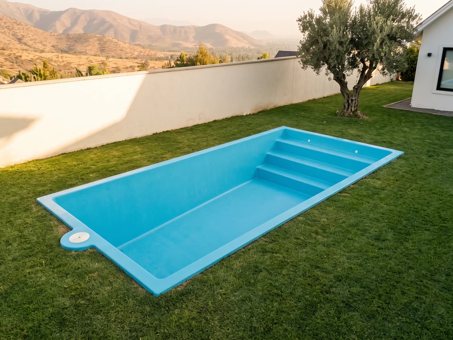 Piscina de fibra de vidrio de 4,50 por 2,50 metros instalada a nivel del pasto en el jardín de una casa, vista desde arriba, vacía y sin borde, con la aleta de fibra a la vista y la escalera de tres peldaños a lo ancho de un extremo