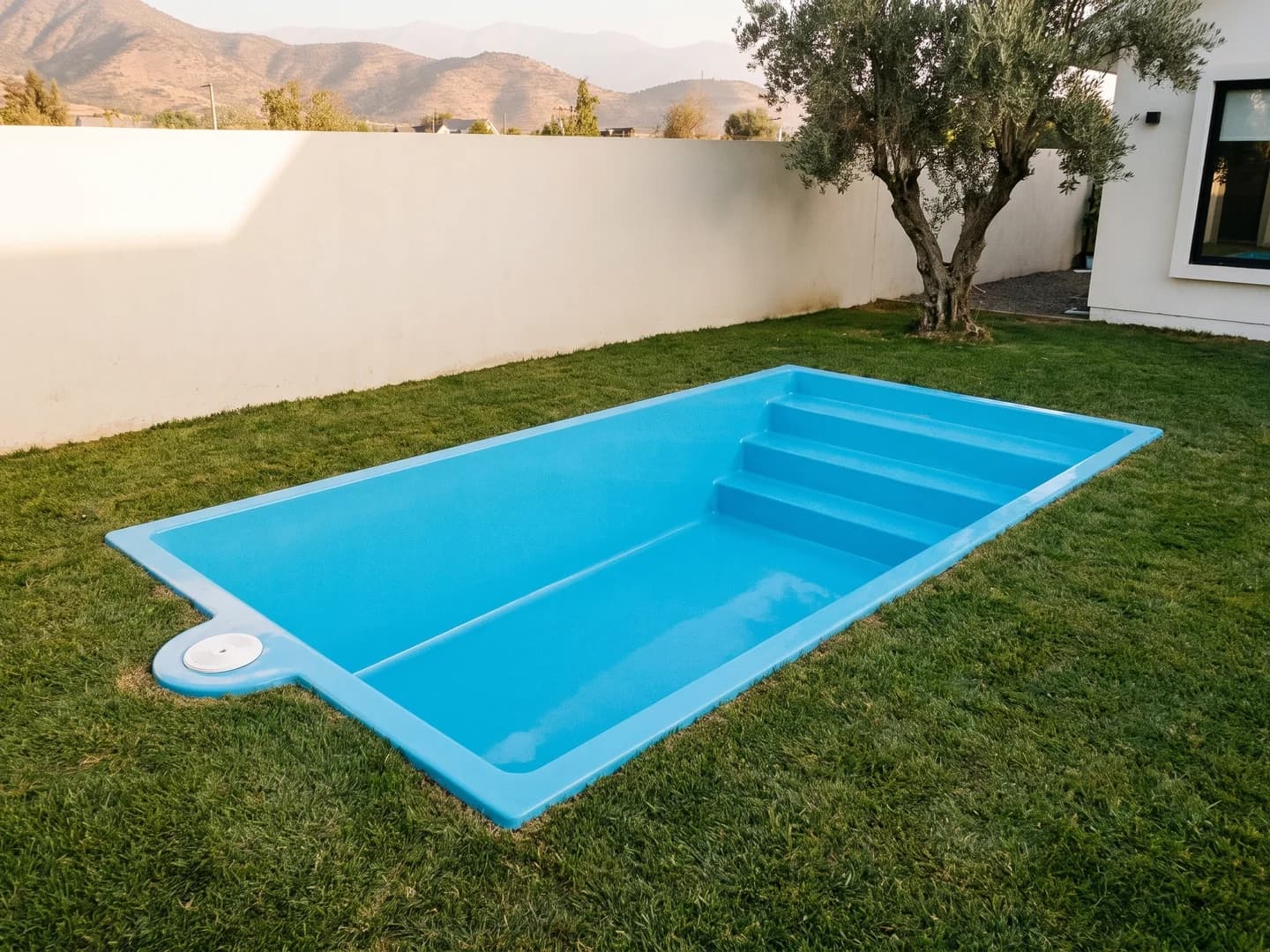 Piscina de fibra de vidrio de 5,00 por 3,00 metros instalada a nivel del pasto en el jardín de una casa, vista desde arriba, vacía y sin borde, con la aleta de fibra a la vista y la escalera de tres peldaños a lo ancho de un extremo
