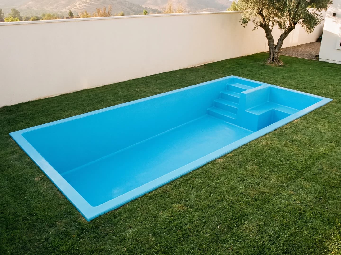 Piscina de fibra de vidrio de 8,40 por 3,40 metros instalada a nivel del pasto en el jardín de una casa, vista desde arriba, vacía y sin borde, con la aleta de fibra a la vista, escalera en una esquina del extremo bajo y una banca en L a su costado